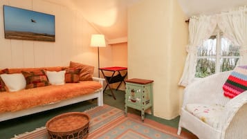 Ein gemütliches Zimmer mit einer Couch, einem kleinen Tisch, einem Korbstuhl, einem Fenster mit Vorhängen und einem Bild an der Wand.