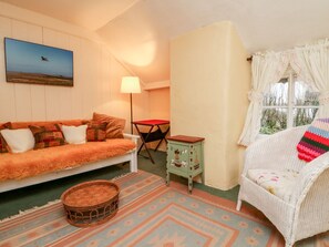 Ein gemütliches Zimmer mit einer Couch, einem kleinen Tisch, einem Korbstuhl, einem Fenster mit Vorhängen und einem Bild an der Wand.