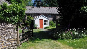 A stone wall, a wooden gate, a small white house with a red door, and a garden with various flowers. - Whitefield Cottage (Barnstaple)