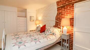 Quarto com uma cama, duas luminárias de cabeceira, uma mesa de cabeceira, um guarda-roupa branco e uma parede de tijolos.