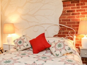 Una cama con ropa de cama floral, un cojín rojo y un cabecero de metal blanco frente a una pared de ladrillo.