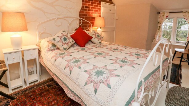 A bedroom with a bed, two bedside tables, a red pillow, a quilt, a brick wall, a window, and a chair. - Whitefield Cottage (Barnstaple)