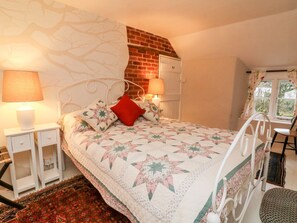 A bedroom with a bed, two bedside tables, a red pillow, a quilt, a brick wall, a window, and a chair. - Whitefield Cottage (Barnstaple)
