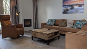 Una sala de estar con una butaca de cuero marrón, una mesa de centro de madera, un sofá marrón con cojines estampados, una chimenea y un cuadro de un atardecer.