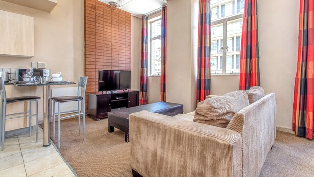 Een woonkamer met een bank, voetenbankje, televisie en een klein keukenhoekje met een tafel en stoelen.