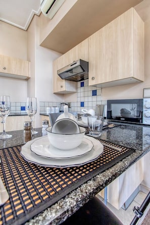 A modern kitchen with light wood cabinets, a stainless steel range hood, a granite countertop, and a dining area with a woven placemat. - 3 Shots Wifi Netflix at Mapungubwe (Johannesburg)