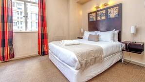 Een hotelkamer met een groot bed, nachtkastje, lamp en een raam met gordijnen.