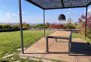 Une terrasse en bois avec une structure en forme de pergola, un luminaire suspendu et un mur de pierre en arrière-plan.