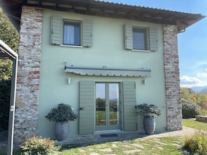 A two-story house with a stone facade, green shutters, and a small porch with potted plants.