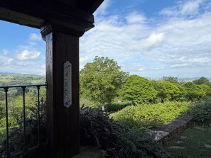 A wooden balcony with a thermometer, lush greenery, and a distant landscape view.