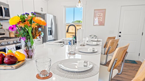 A modern kitchen with white cabinetry, a central island with a vase of flowers, and a dining area with chairs and a table set for two. - NEW BUILD | 10 Mins to Folly | 4 BR, 10 Guests (Charleston)