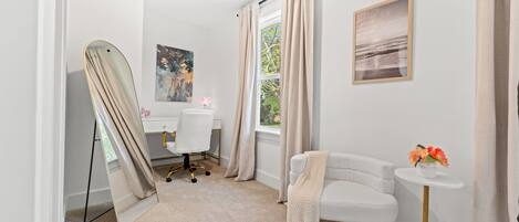 Une pièce avec un grand miroir, un bureau, une chaise, un canapé, une petite table ornée de fleurs et une fenêtre avec des rideaux.