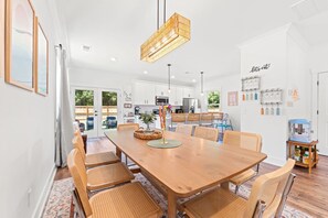 A modern kitchen with white cabinets, a dining table with wicker chairs, and a hanging light fixture. - NEW BUILD | 10 Mins to Folly | 4 BR, 10 Guests (Charleston)