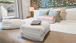 A neatly made bed with folded towels, a headboard with floral wallpaper, and a view of houses through a window. - NEW BUILD | 10 Mins to Folly | 4 BR, 10 Guests (Charleston)