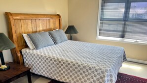 Un letto con copriletto decorato, una testiera in legno, due abat-jour, un tappeto rosso con motivi e una finestra con persiane.