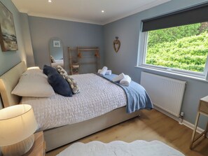 A bedroom with a bed, a bedside table with a lamp, a window with a view of greenery, and a radiator. - Tigh-na-mara Retreat (Berwick-upon-Tweed)