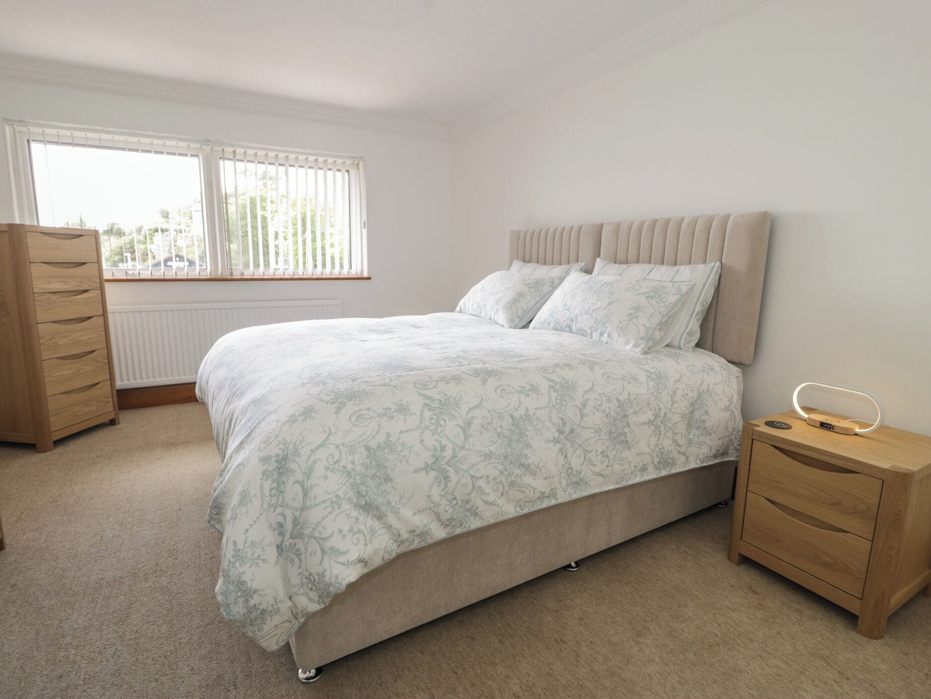 A bedroom with a bed, a dresser, a bedside table with a lamp, and a window with blinds.
