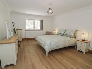 Ein Schlafzimmer mit Holzboden, einem Bett mit floralem Bettbezug, einem Nachttisch mit Lampe, einem Schminktisch und einem Fenster mit Vorhängen.