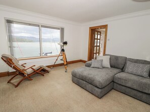 A living room with a view of the sea, a wooden rocking chair, a gray sofa, and a telescope. - The Boat Yard Apartment (Porthmadog)
