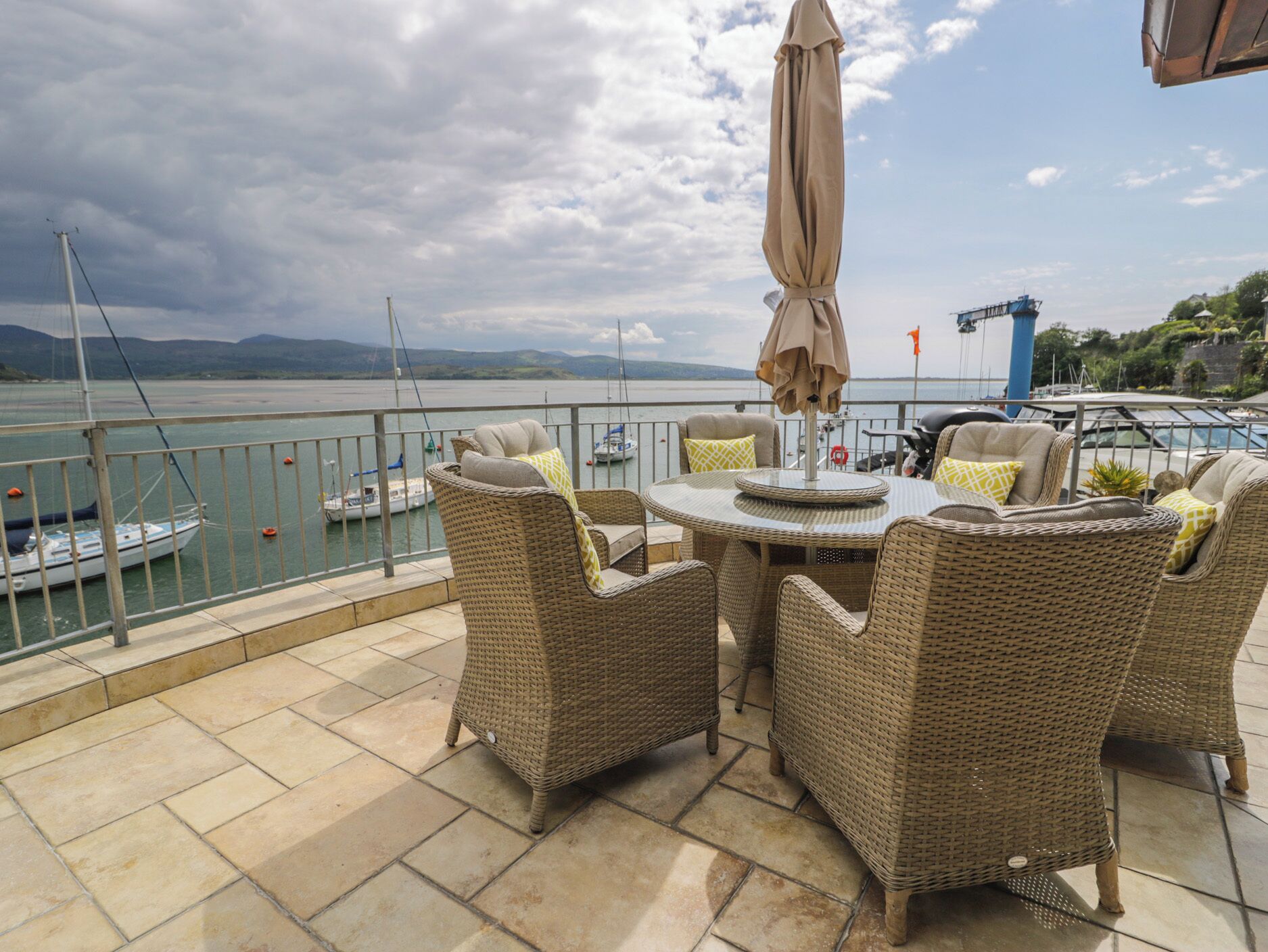 A terrace with wicker furniture and an umbrella, overlooking a marina with boats and a cloudy sky.