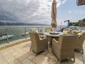 A terrace with wicker furniture and an umbrella, overlooking a marina with boats and a cloudy sky. - The Boat Yard Apartment (Porthmadog)