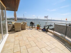 Eine Terrasse mit Korbmöbeln, einem Grill, Topfpflanzen und Blick auf das Wasser.