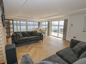 A spacious living area with a large gray sofa, a dining table, and a view of the sea through large windows. - The Boat Yard Apartment (Porthmadog)
