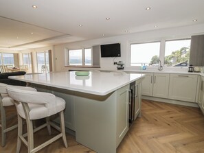 A modern kitchen with a large island, bar stools, a flat-screen TV, and a view of the outdoors. - The Boat Yard Apartment (Porthmadog)