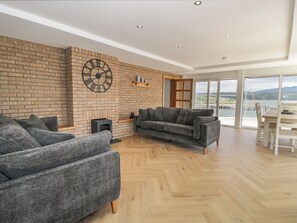 A spacious living room with a brick wall, a large clock, and a view of a lake through large windows. - The Boat Yard Apartment (Porthmadog)