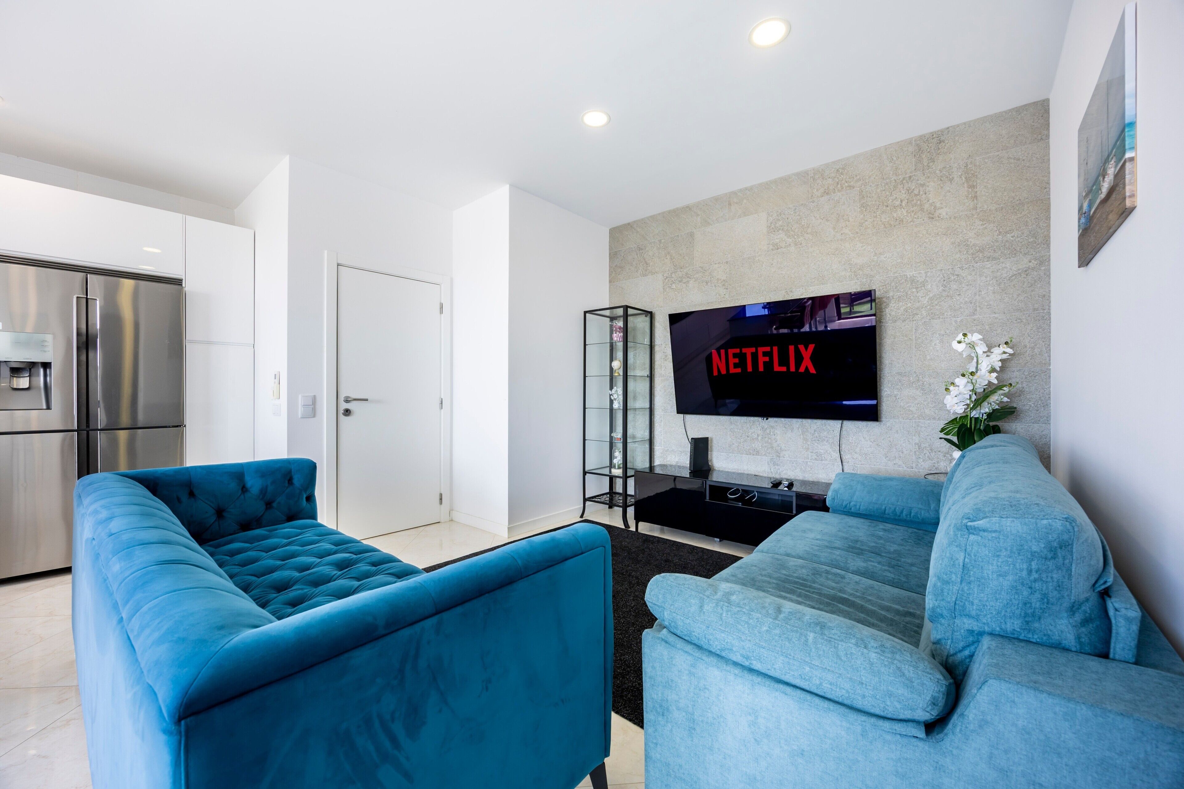 Een moderne woonkamer met twee blauwe banken, een flatscreen televisie bevestigd aan een structuurwand, een glazen kast en een keuken met roestvrijstalen apparatuur.