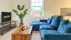 A living room with a TV, a blue sofa, a wooden coffee table, a large plant, and a window with a view of buildings. - Cosy & Stylish Canterbury Apartment | Free Parking (Canterbury)