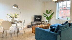 A modern living room with a blue sofa, a dining table with chairs, a TV on a stand, and a large window with a view of buildings. - Cosy & Stylish Canterbury Apartment | Free Parking (Canterbury)