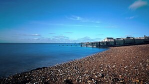A pebbled beach with a pier extending into calm waters, a clear sky, and a distant building. - Cosy & Stylish Canterbury Apartment | Free Parking (Canterbury)