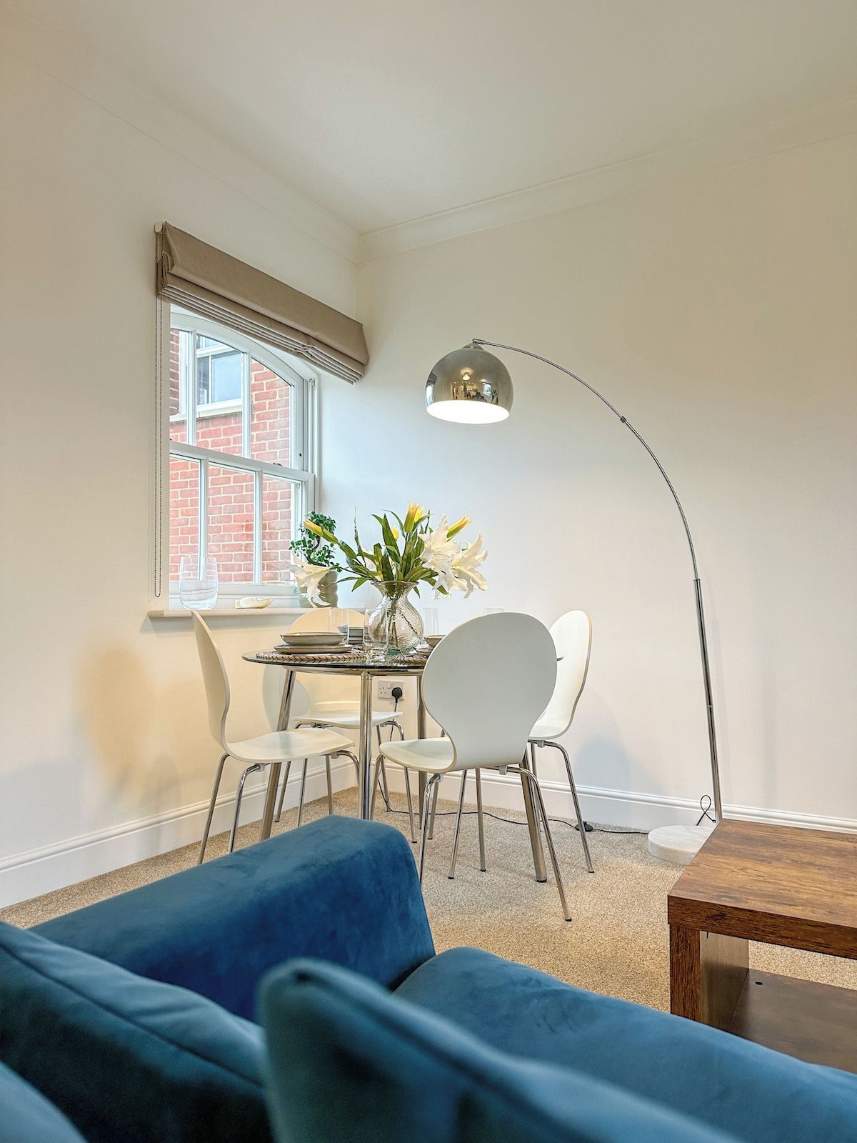 Um quarto com mesa de jantar, duas cadeiras, um vaso com flores, um sofá azul e um abajur.