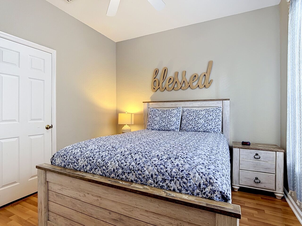 Une chambre à coucher avec un lit, une table de chevet, une commode, une lampe et un mur orné d’une inscription « béni ».