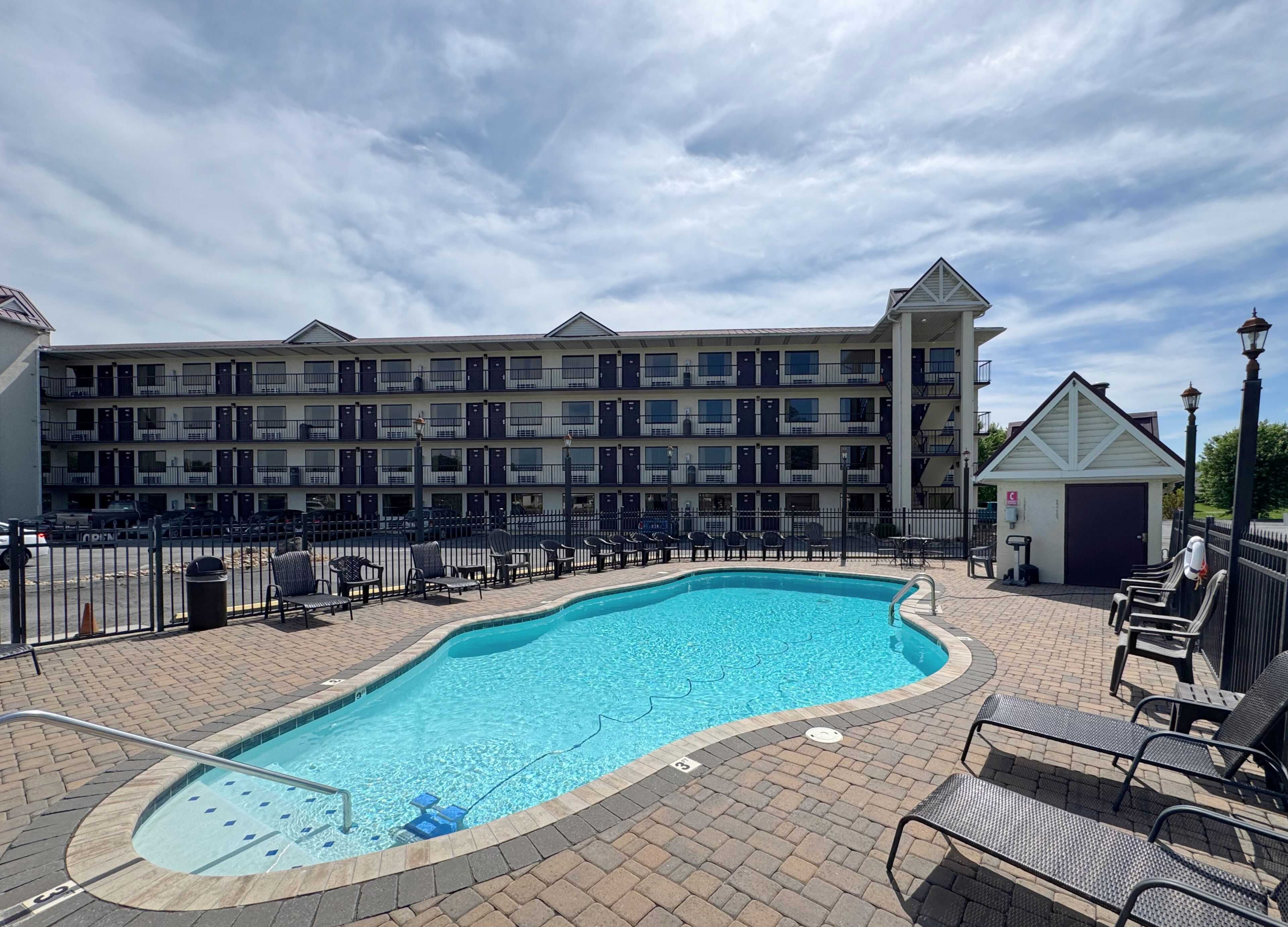 A swimming pool with lounge chairs, a small building, and a multi-story hotel in the background.