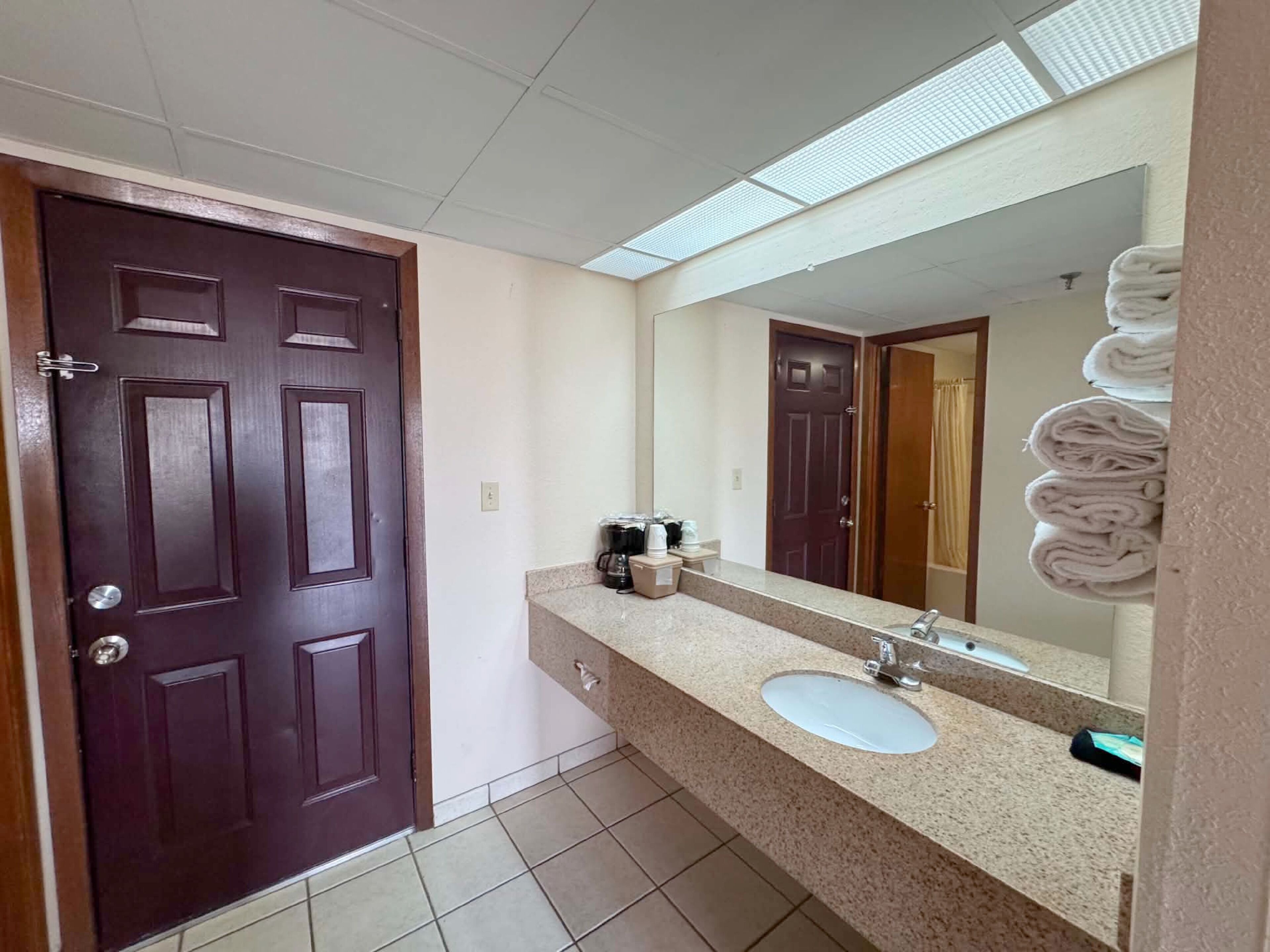A bathroom with a dark wooden door, a white sink, a large mirror, and a towel rack with folded towels.