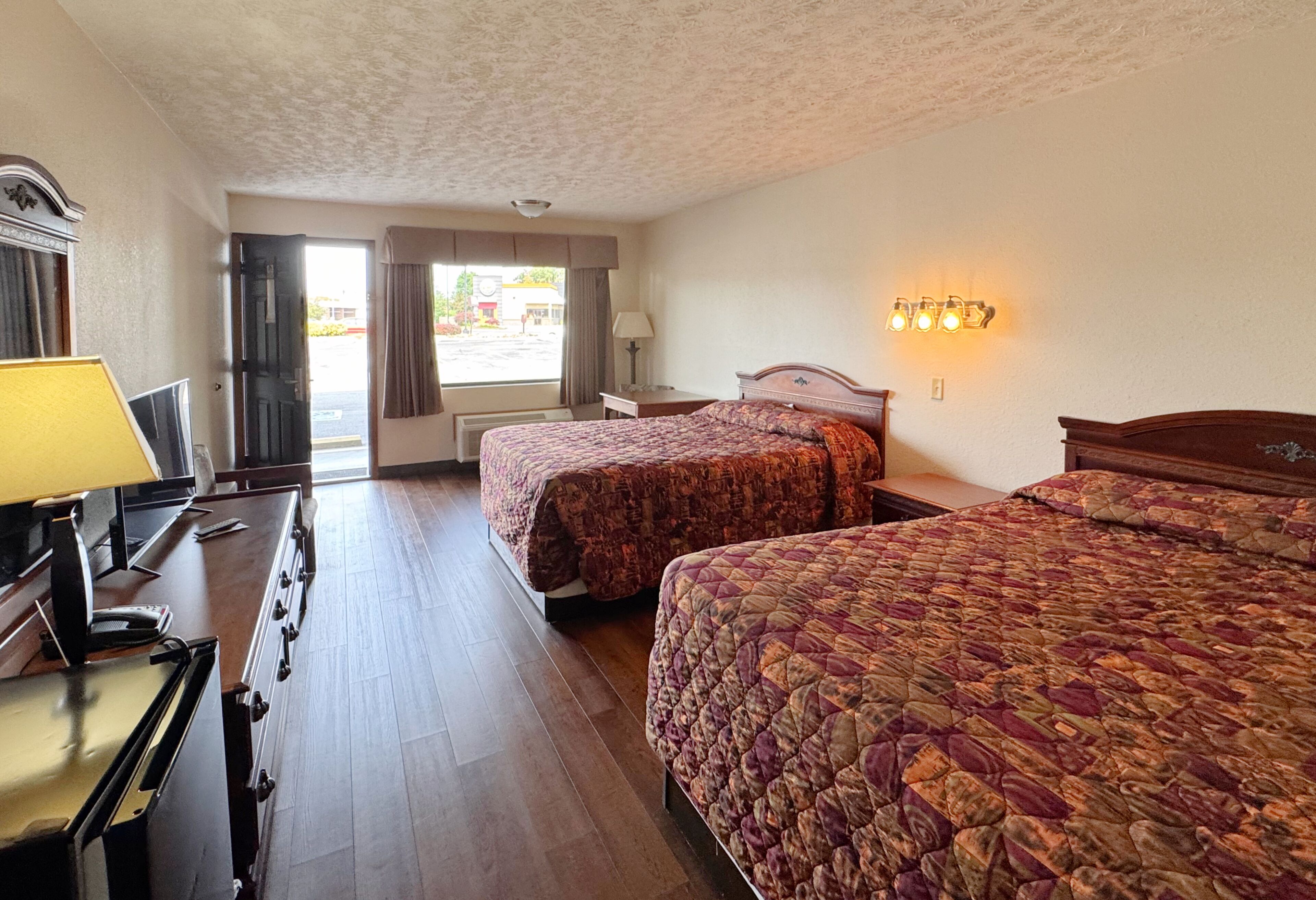A hotel room with two beds, a desk with a lamp, a dresser, and a view of the street.