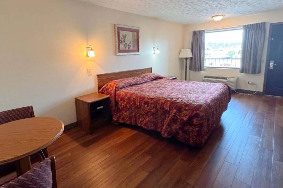 A bedroom with a bed, a bedside table, a chair, a table, a window with curtains, and a wall-mounted picture.