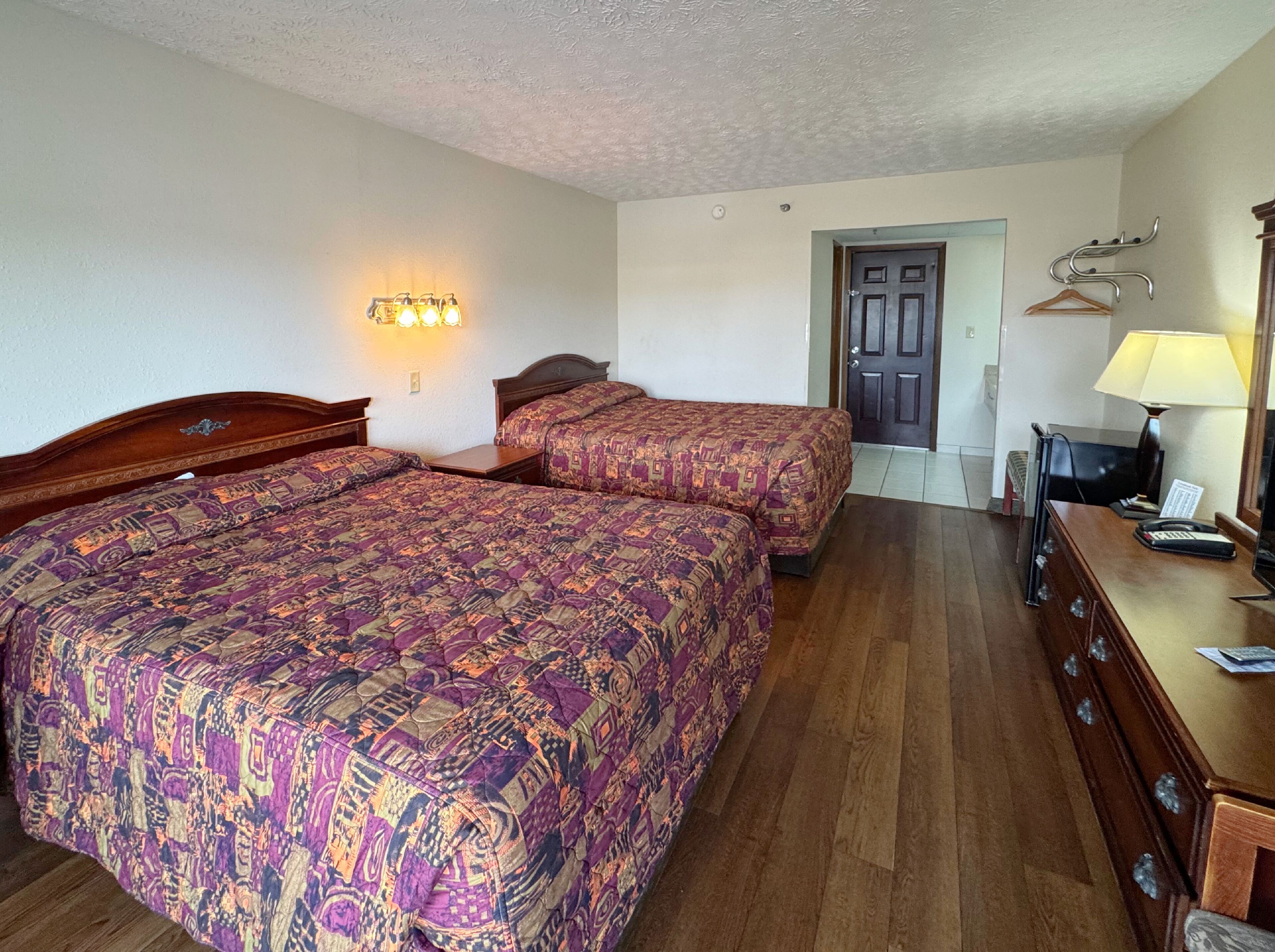 Quarto de hotel com duas camas, uma escrivaninha com abajur, um cabideiro e uma porta.
