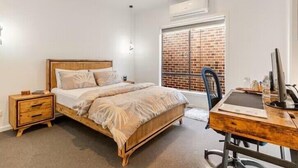 Une chambre à coucher avec un lit en bois, un bureau avec un ordinateur, une chaise, une table de chevet et une fenêtre donnant sur un mur de briques.