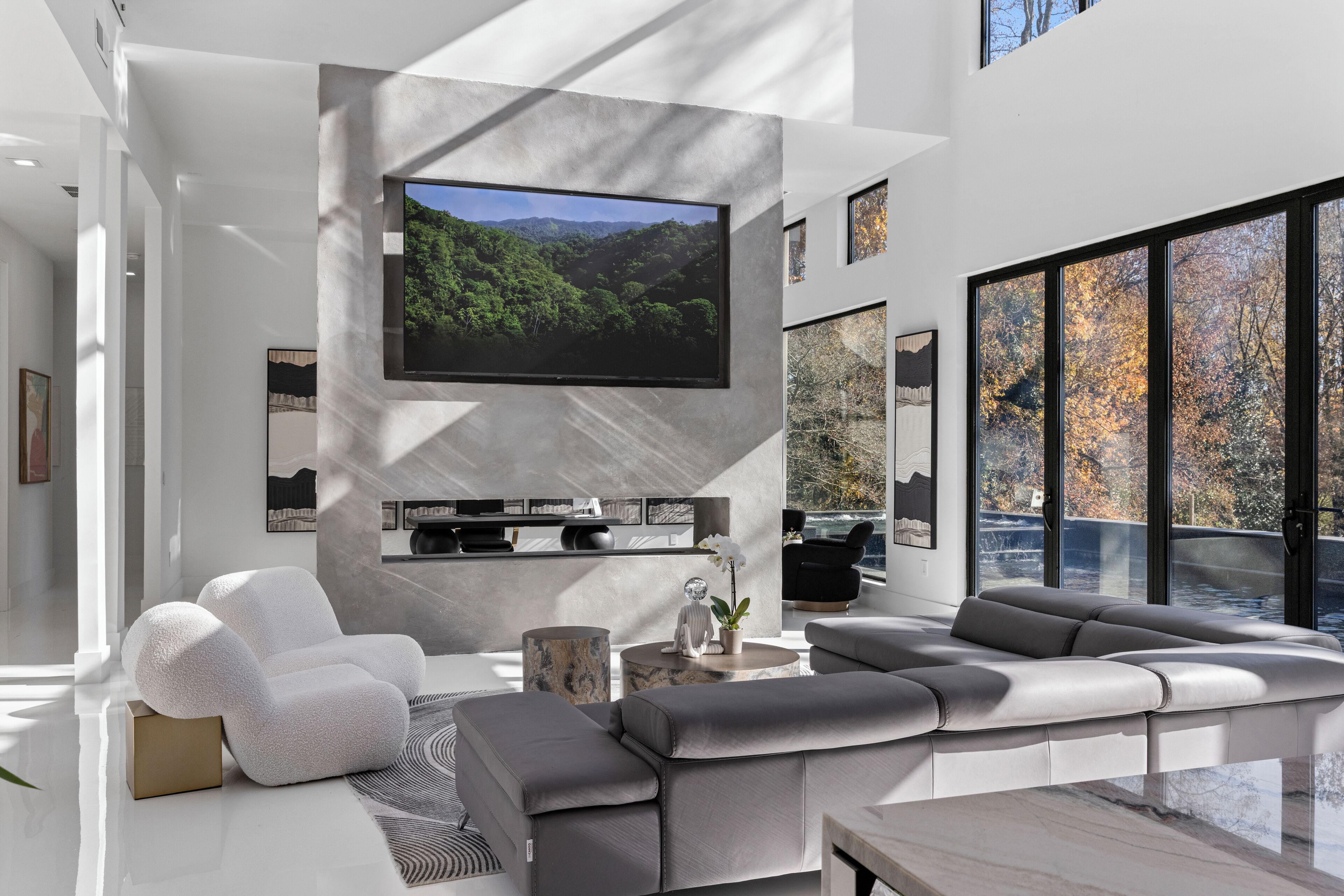 A modern living room with a large flat-screen TV, a grey sectional sofa, a white armchair, and a coffee table.