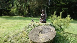 A statue in a garden with a tree stump, grass, and pine trees in the background.