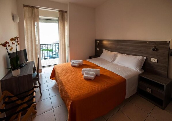 A hotel room with a bed, a desk with a chair, a TV, and a balcony with curtains.