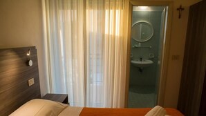 Ein Hotelzimmer mit Bett, Nachttisch, Bad mit Waschbecken und Spiegel sowie einem Kruzifix an der Wand.