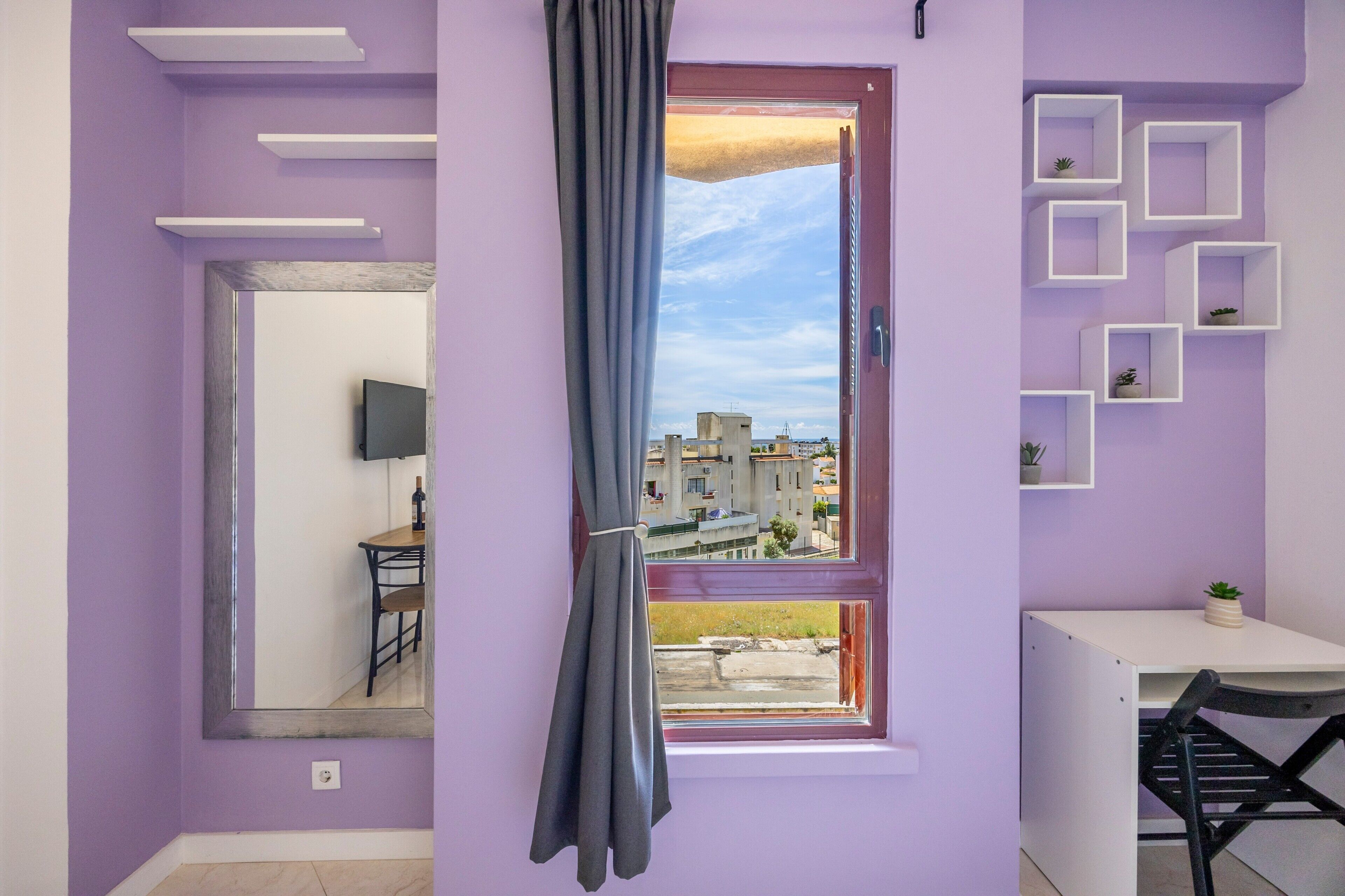 A room with a purple wall, a window with a view of buildings, a mirror, a small table, and a chair.