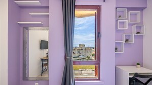 Ein Zimmer mit violettem Wand, einem Fenster mit Blick auf Gebäude, einem Spiegel, einem kleinen Tisch und einem Stuhl.