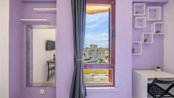 A room with a purple wall, a window with a view of buildings, a mirror, a small table, and a chair.