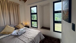Un dormitorio con cama, cortinas, una mesita de noche, una lámpara y dos ventanas con vistas al verdor.