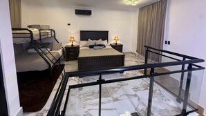 Habitación de hotel con cama, escritorio, silla, ventana con cortinas, lámpara en el techo y escalera con barandilla de vidrio.